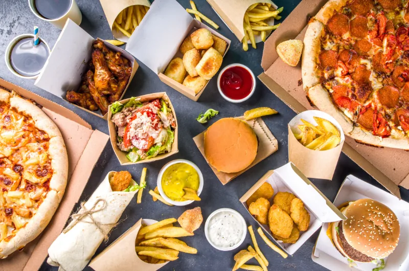 Vue d'ensemble d'un repas fast-food viral : burgers, pizzas, wings, nuggets, frites et sauces partagés en boîtes carton.